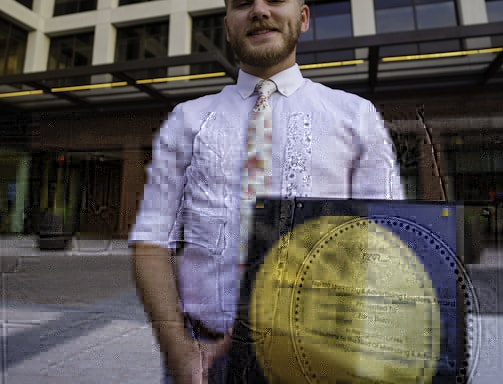 A person in a white shirt with a floral tie is holding a large square plaque with a round gold emblem. The plaque is an award for marketing and advertising, and the person is standing in front of a modern office building with large windows.