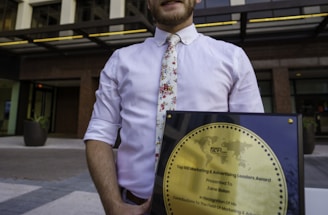 A person in a white shirt with a floral tie is holding a large square plaque with a round gold emblem. The plaque is an award for marketing and advertising, and the person is standing in front of a modern office building with large windows.