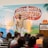 A man is standing on a stage in front of a backdrop with the text 'Social Media Marketing World'. He is wearing a patterned shirt and white pants, gesturing as he speaks. There is a podium with a microphone and two books on it. An audience is seated and listening attentively.