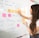 A woman with long hair is writing on a whiteboard. She is using sticky notes and markers to organize and plan a strategy. The board has various handwritten notes and symbols related to marketing and digital education.